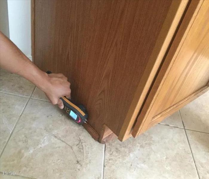 A close up of a tile floor and the corner of a cabinet with a persons hand using a moisture meter to check if its dry