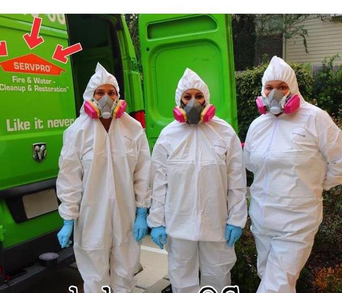 3 women in white Tyvek suits and blue gloves standing for a picture in front of a green SERVPRO van